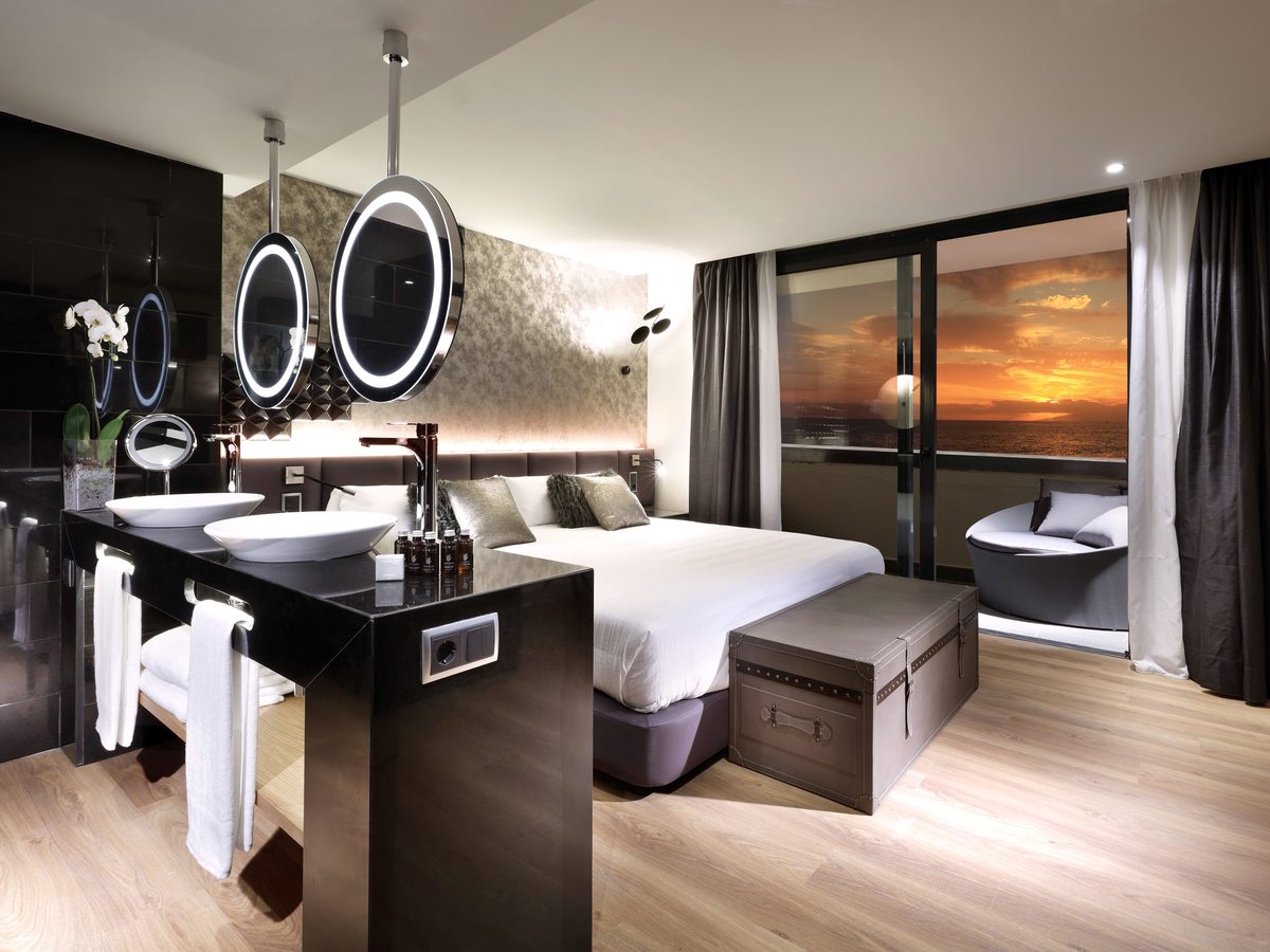 Open-plan suite with a freestanding bath and twin illuminated mirrors beside the bed, and a sea-view balcony.
