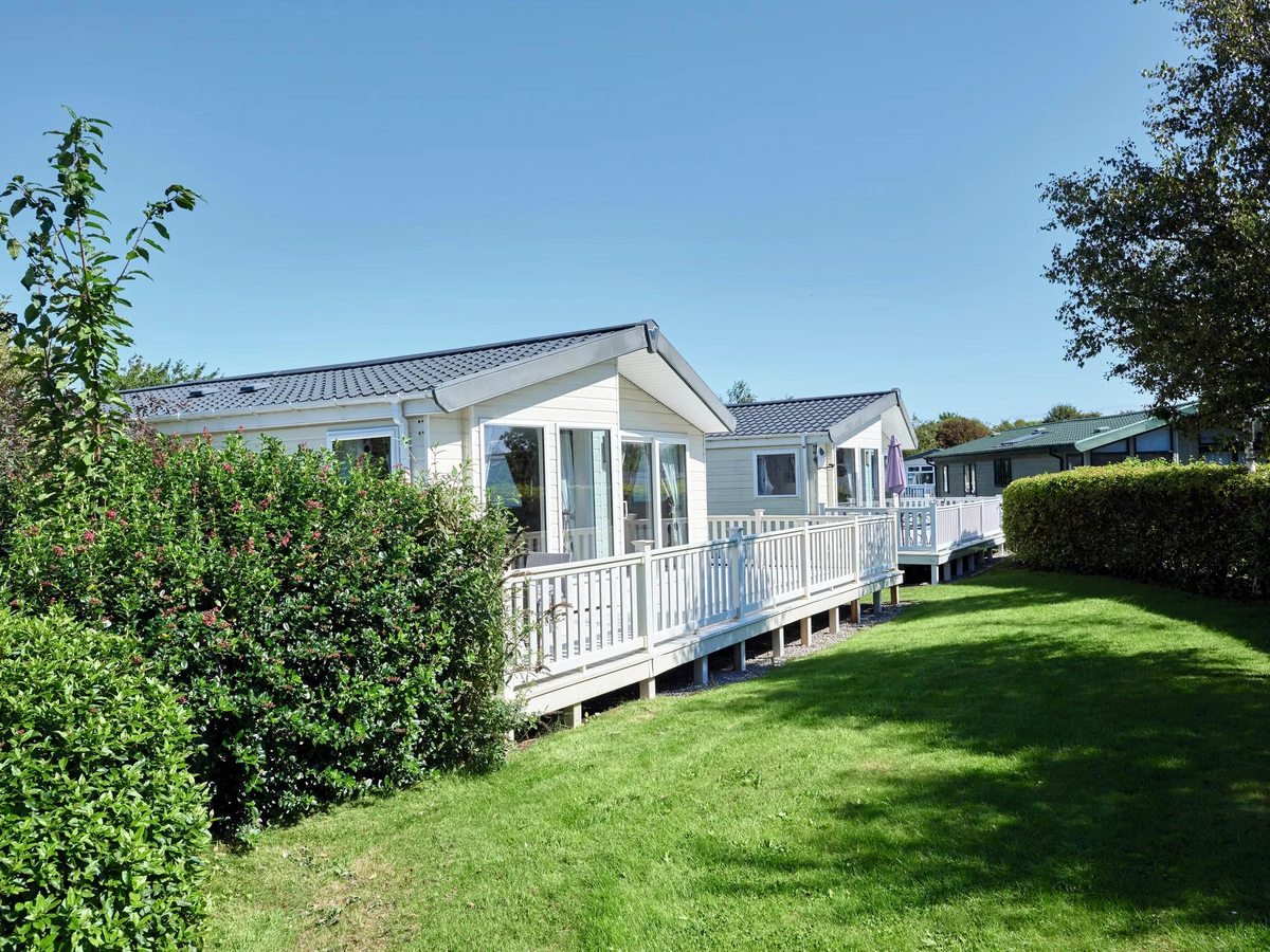 A pale-grey holiday home with a white-railed veranda on a bright green lawn under a clear sky.