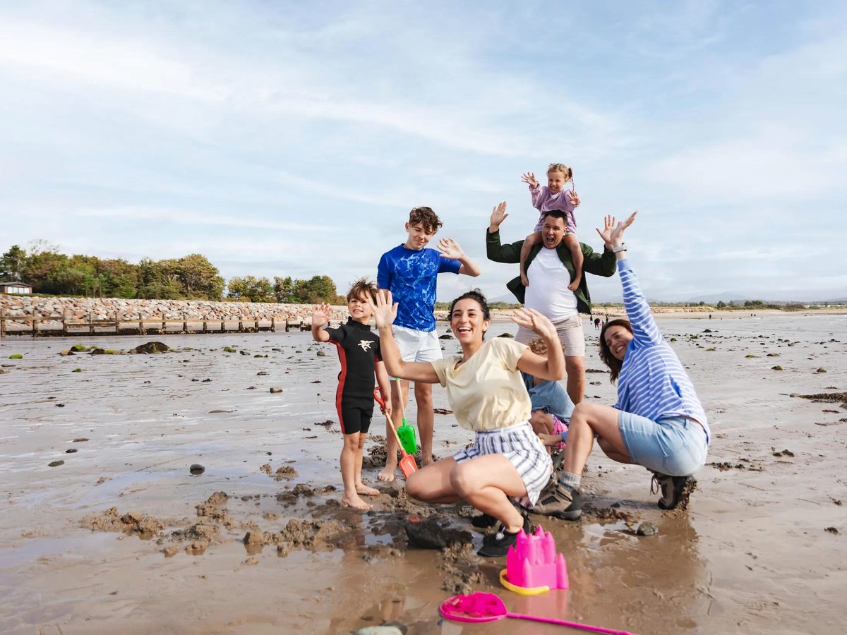 A family on the wide muddy-sand beach at low tide, a rock groyne and distant hills behind.