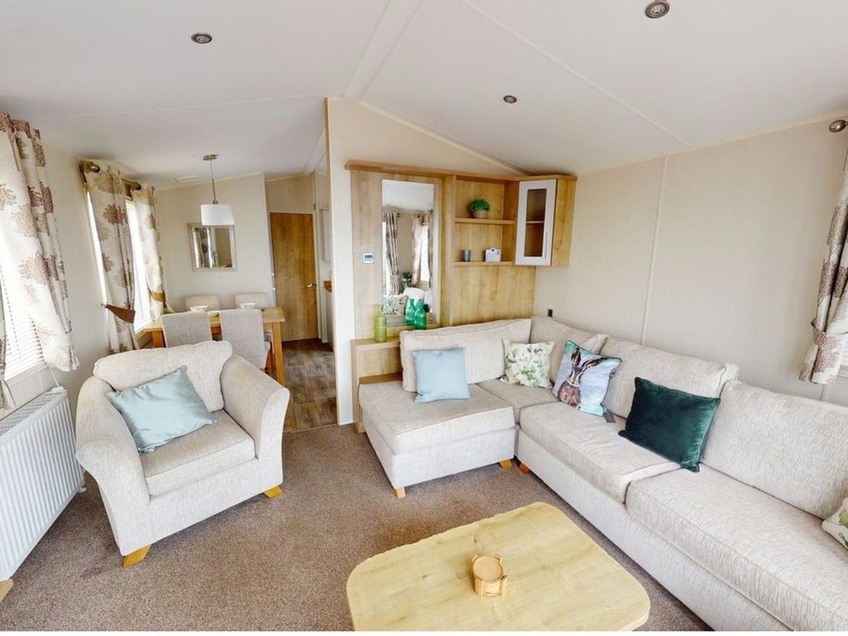 An open-plan caravan lounge with a cream corner sofa, an armchair, an electric fire and a dining area beyond.