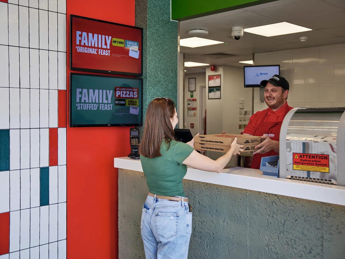 A member of staff handing pizza boxes to a customer at the Papa John's counter, menu screens above.
