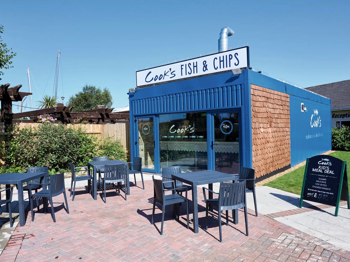 The blue Cook's Fish and Chips unit with outdoor tables and a kid's meal deal board, boat masts behind.
