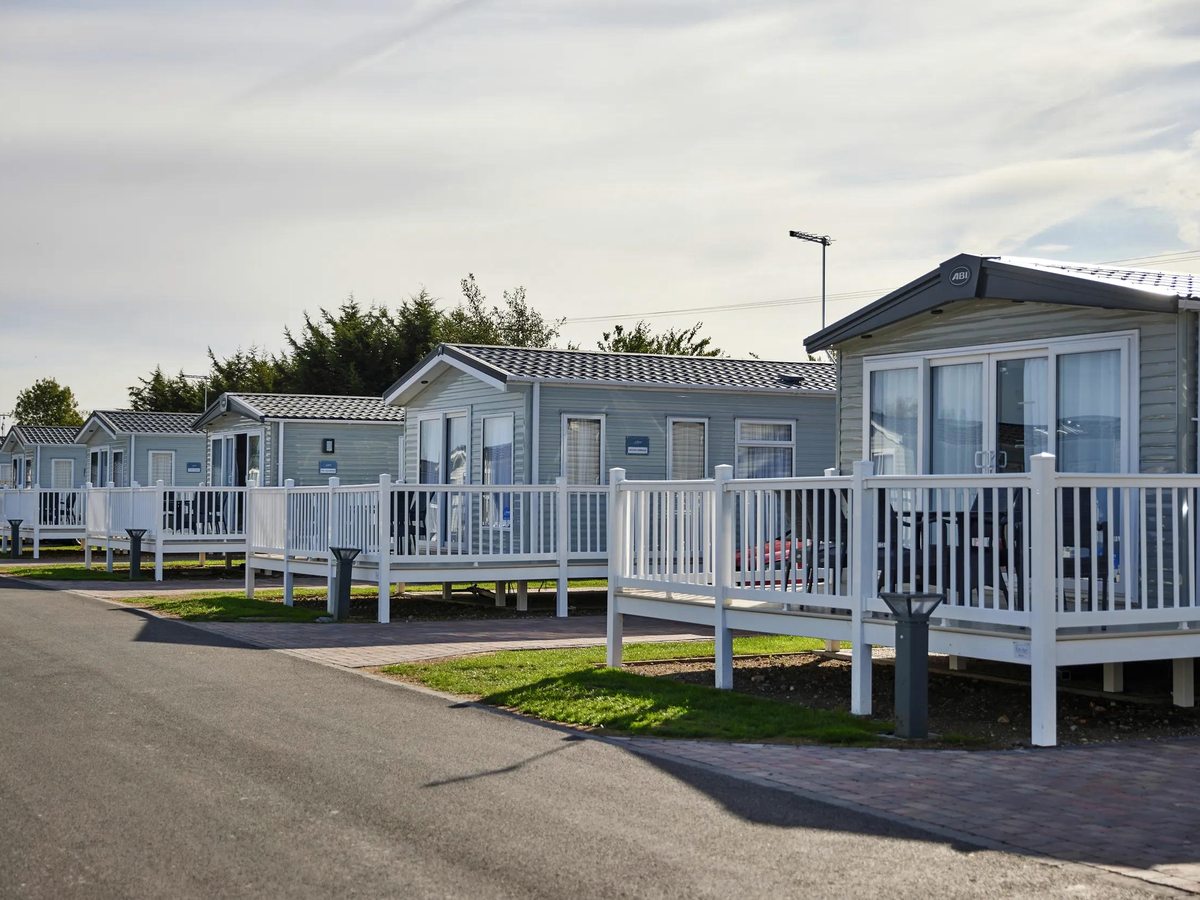White caravans with picket-fenced verandas lined along a tarmac road, a conifer hedge behind.