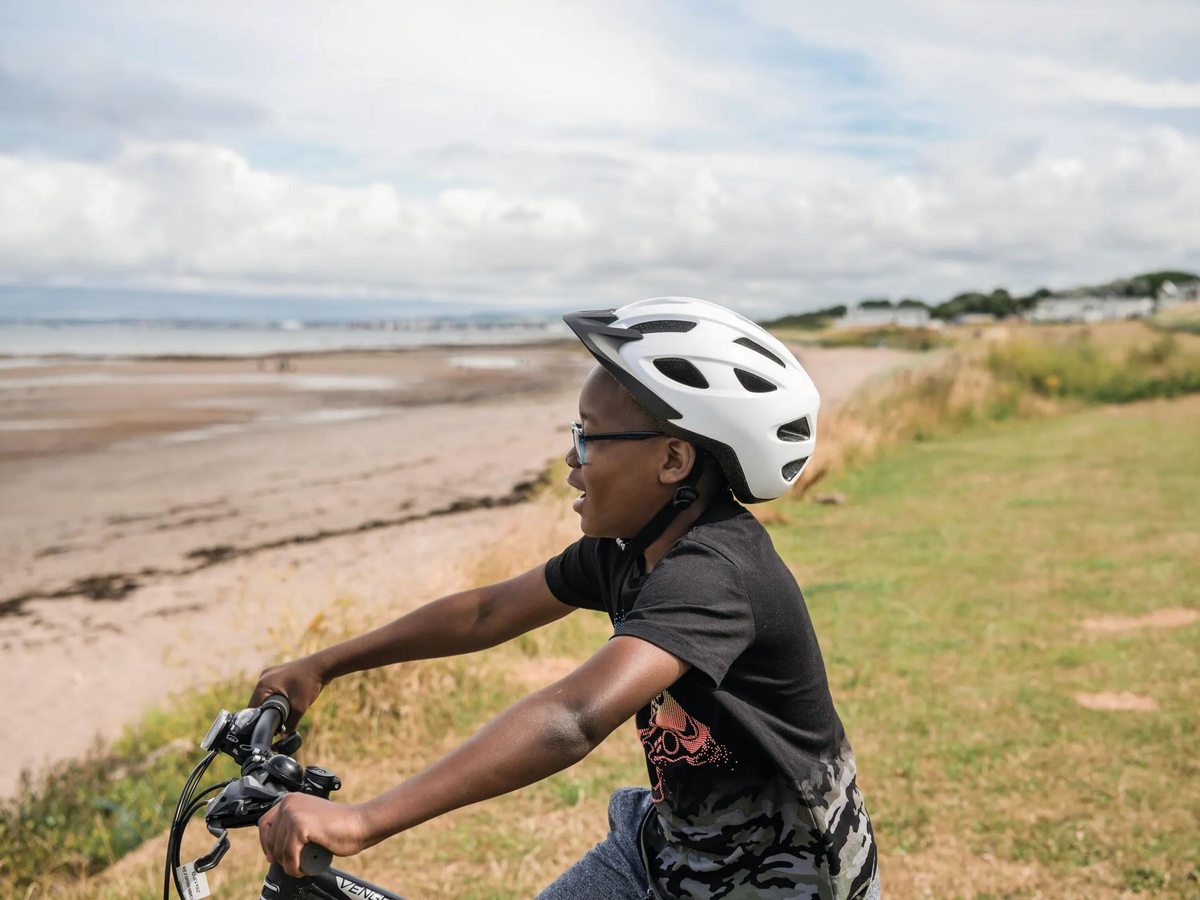 A boy cycling a grassy coastal path beside the wide low-tide beach, the town and hills in the distance.