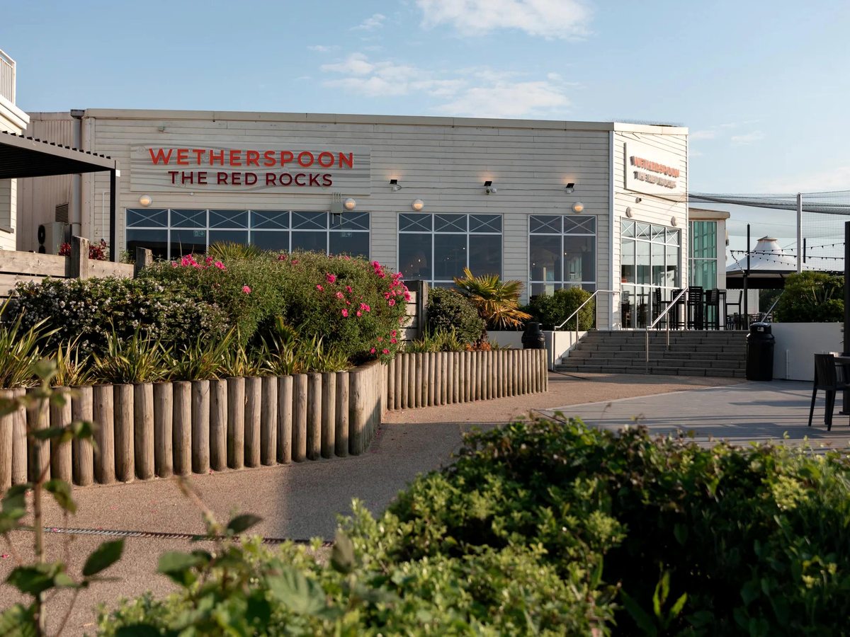The exterior of The Red Rocks at Haven Devon Cliffs, a cream-clad Wetherspoon pub with a red sign reading Wetherspoon The Red Rocks, flower beds and steps up to the entrance in warm evening light.
