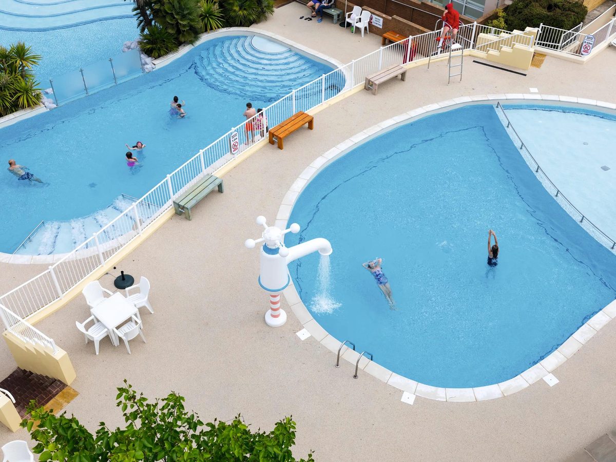 An aerial view of two curved outdoor pools at Haven Devon Cliffs, edged with pale paving, sun loungers and a lifeguard chair, with a few families swimming.