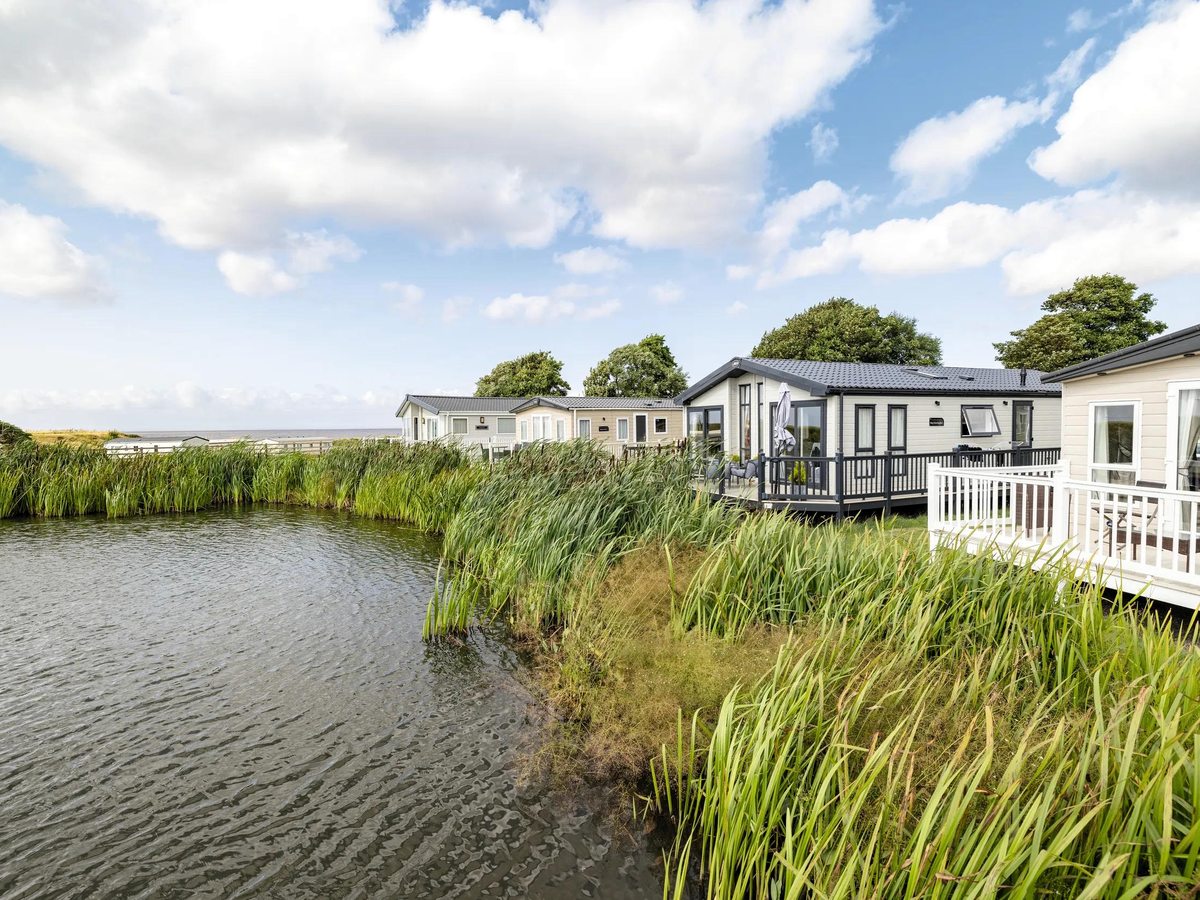 Modern grey holiday caravans and lodges with decking beside a reed-fringed lake at Haven Devon Cliffs, with the sea on the horizon.