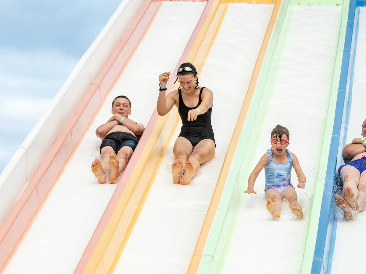 Adults and children racing down side-by-side pastel-coloured mat waterslides against a blue sky.