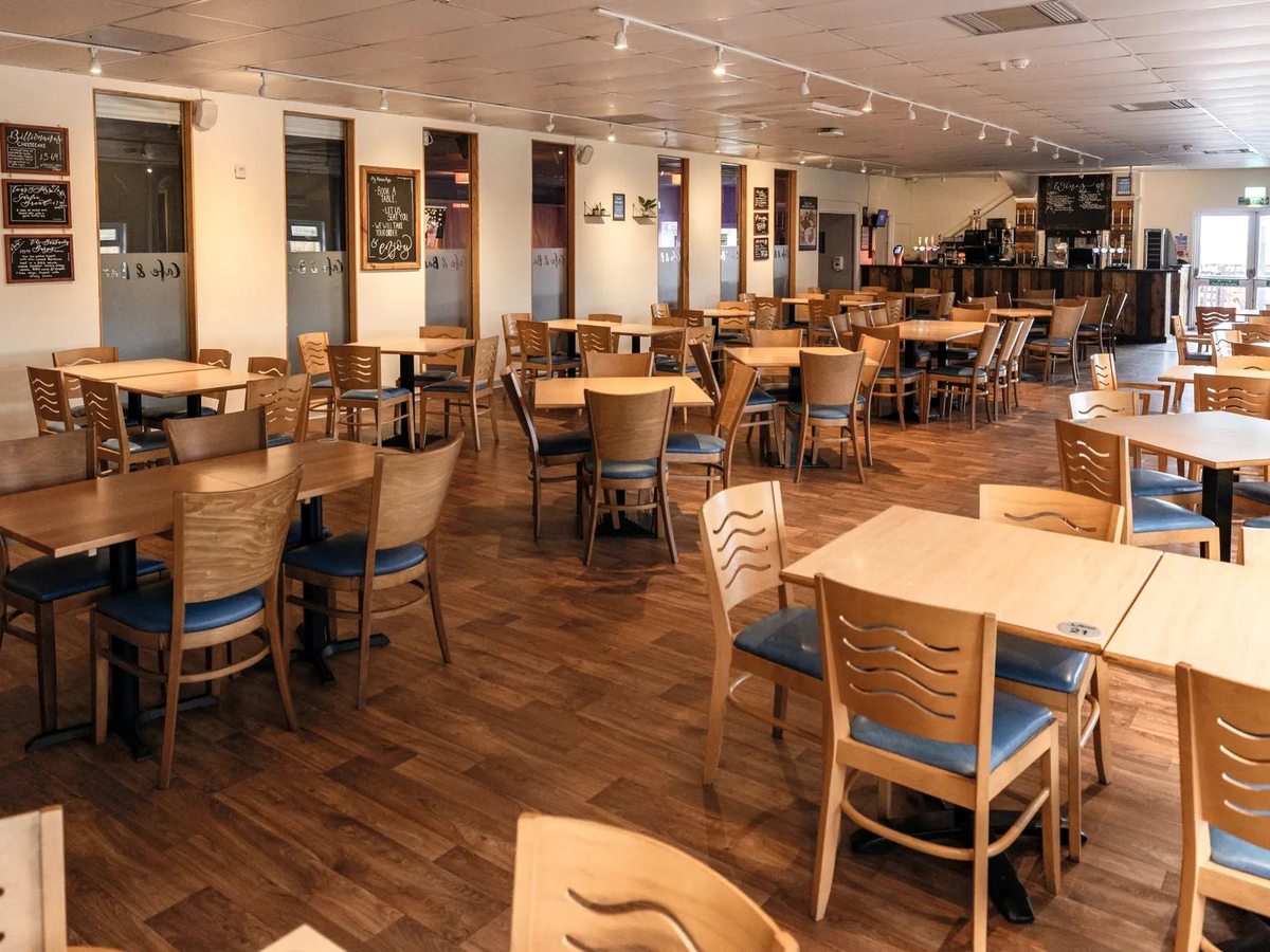 The interior of a cafe-bar restaurant at Haven Devon Cliffs, with light wooden tables and chairs on a wood-effect floor, chalkboard menus on the walls and a bar counter at the far end.