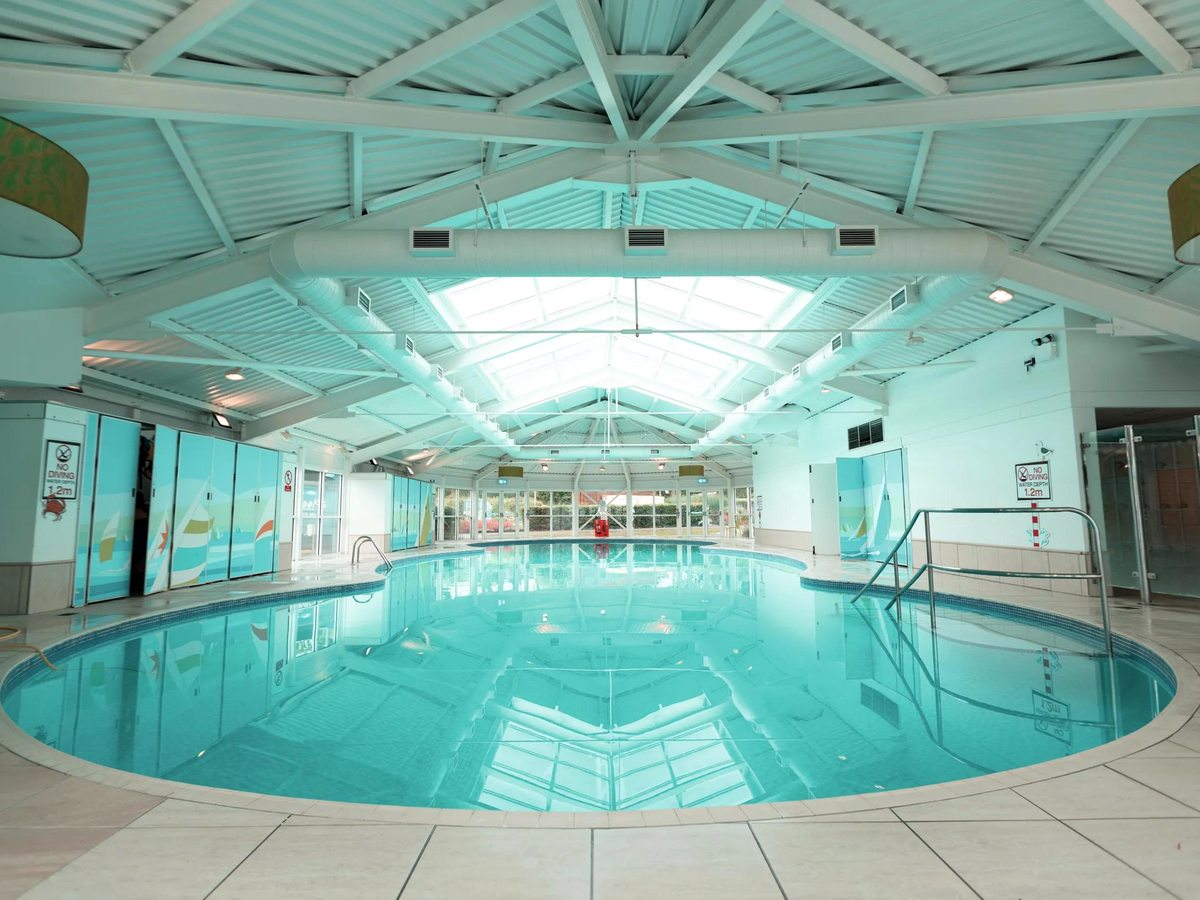 The indoor pool at Haven Devon Cliffs, a curved turquoise pool under a white pitched roof with a glazed apex, shallow steps and handrails at the near end.