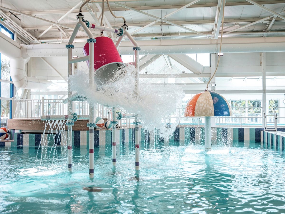 A large bucket tipping water over the play area of the indoor pool, with a raised walkway behind.