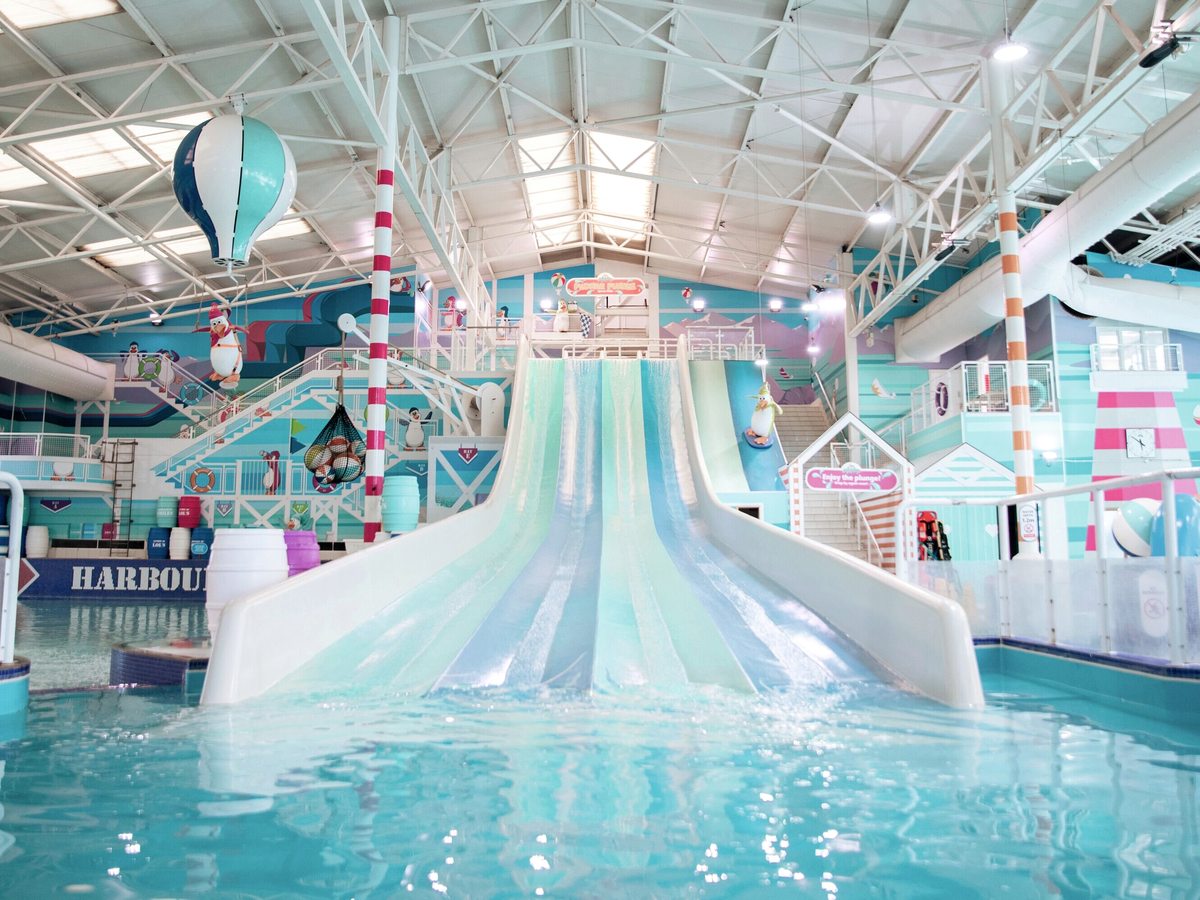 White flumes and a multi-lane racer slide rising above the pool inside the indoor waterpark, under a glazed roof.