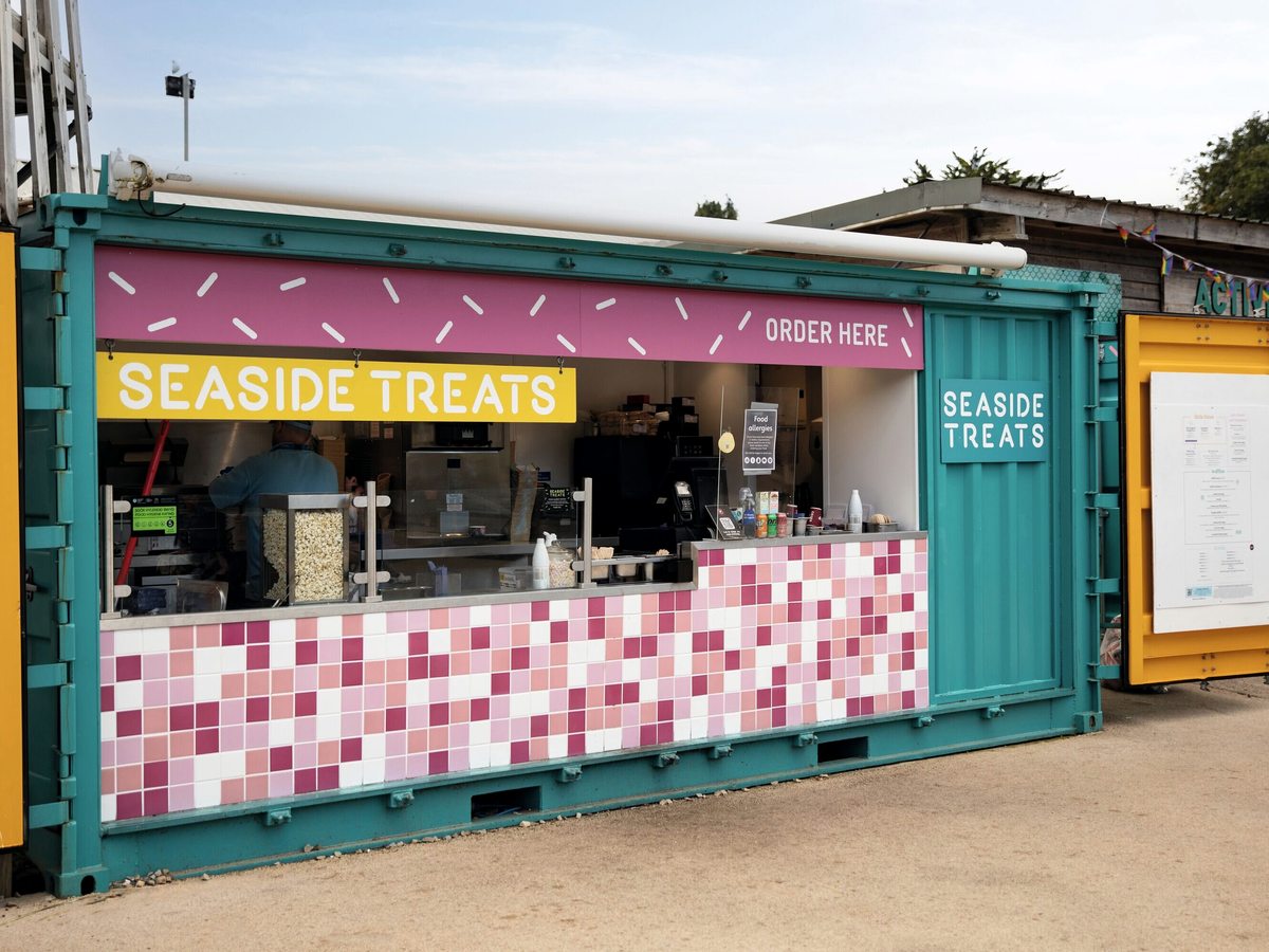 A turquoise shipping-container kiosk with a pink-and-white tiled counter and a Seaside Treats sign over the order window.