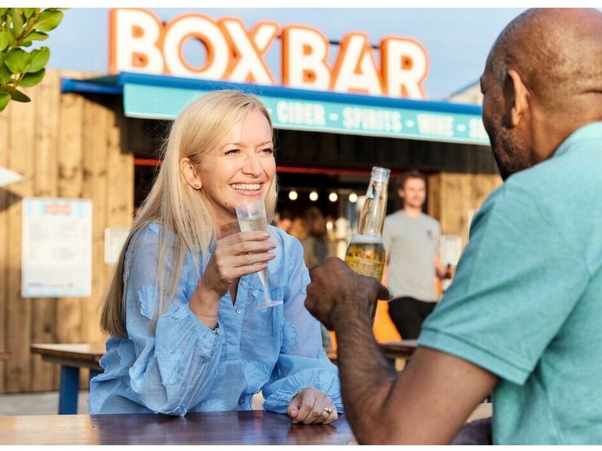 A timber-clad outdoor bar kiosk with a Box Bar sign and a couple sitting with drinks in front.
