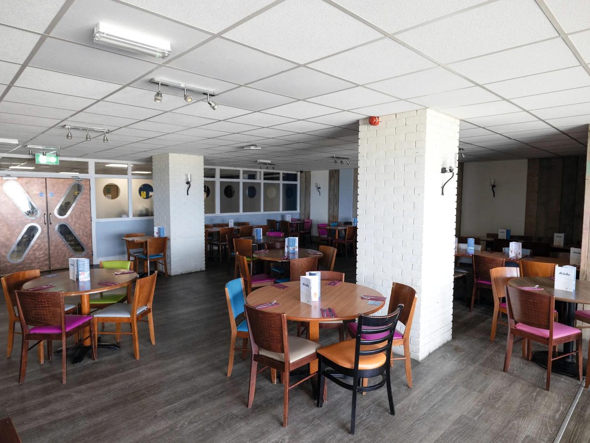 An indoor dining room with round wooden tables and coloured chairs on a grey wood-effect floor.
