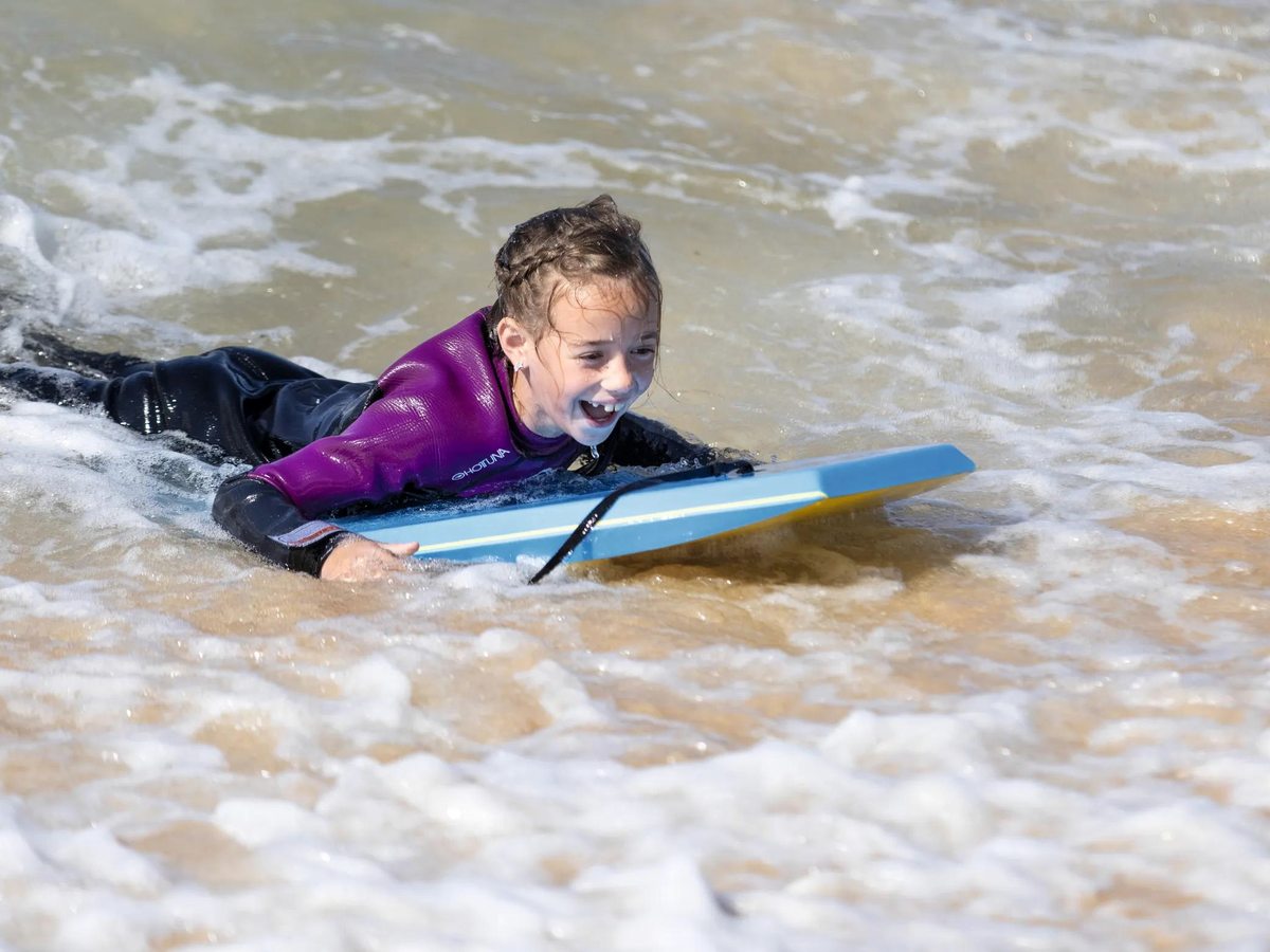 A young girl in a wetsuit lying on a bodyboard, laughing as she rides white surf.