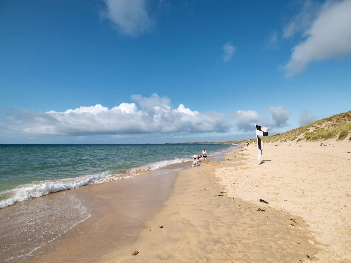 A wide pale-sand beach backed by dunes with gentle waves and a headland in the distance under a blue sky.