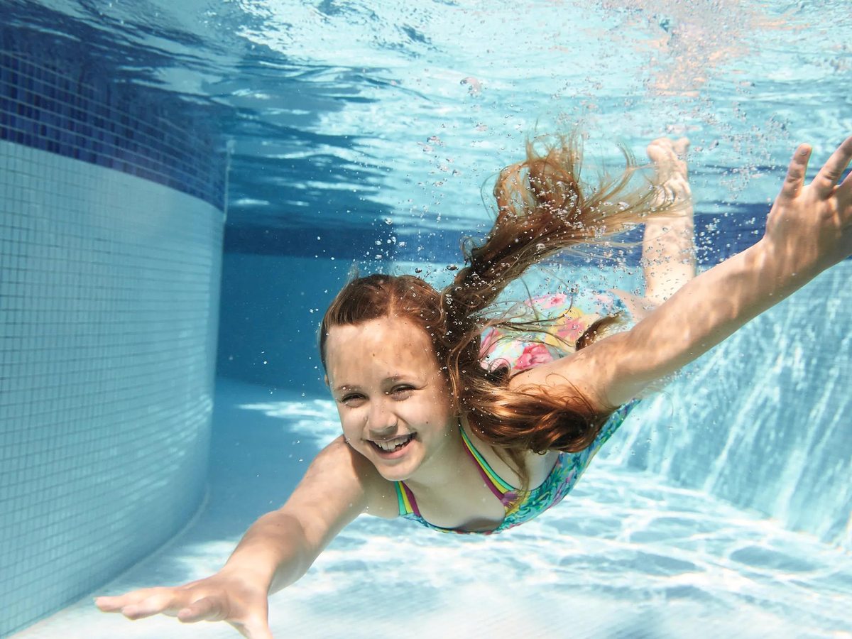 A young girl in a swimsuit swimming underwater in a clear blue pool, smiling.