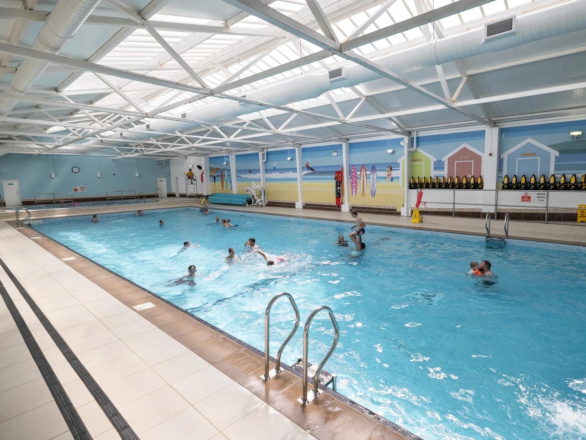 A rectangular indoor swimming pool with people swimming, a pitched roof with skylights and a painted beach-hut mural along one wall.