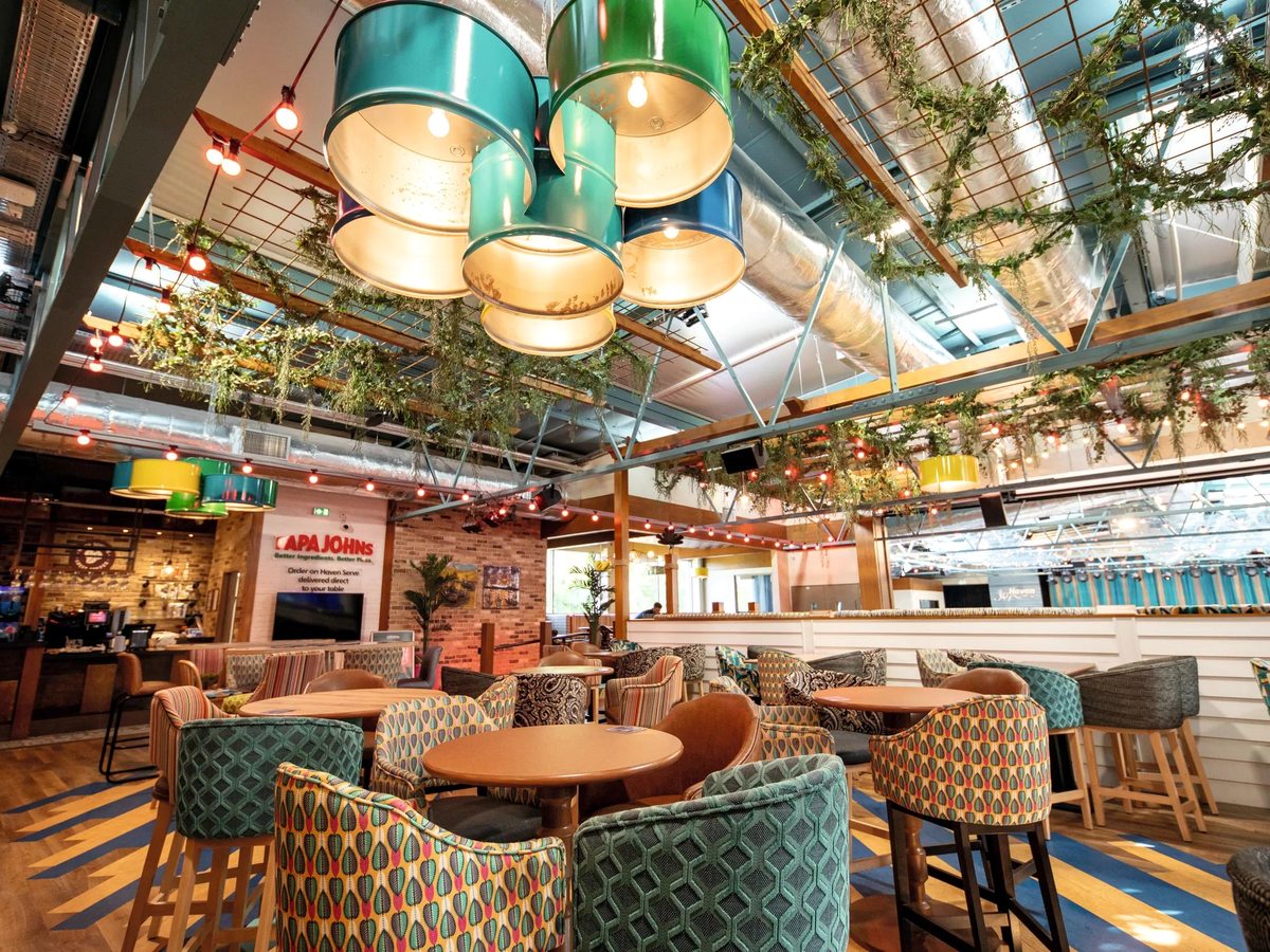 An indoor lounge with patterned tub chairs, hanging drum lampshades and a Papa Johns counter in the background.