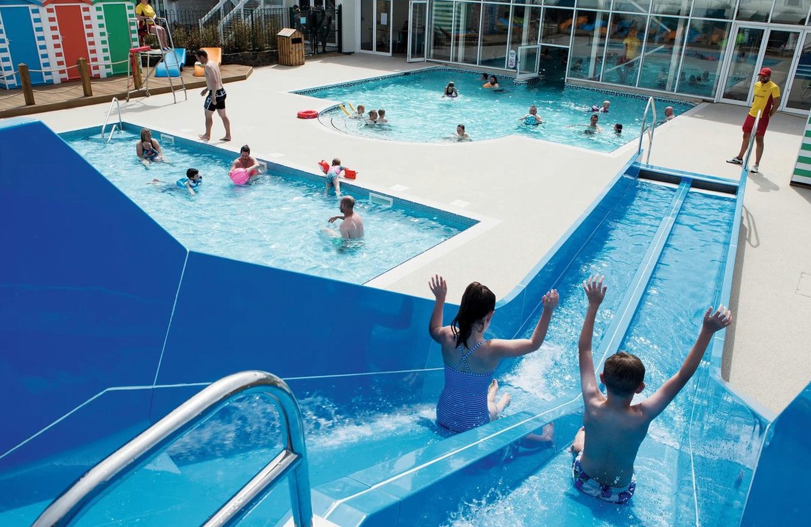 The outdoor pool at Haven Riviere Sands, with families in the water, a two-lane slide and the indoor pool building behind on a sunny day.