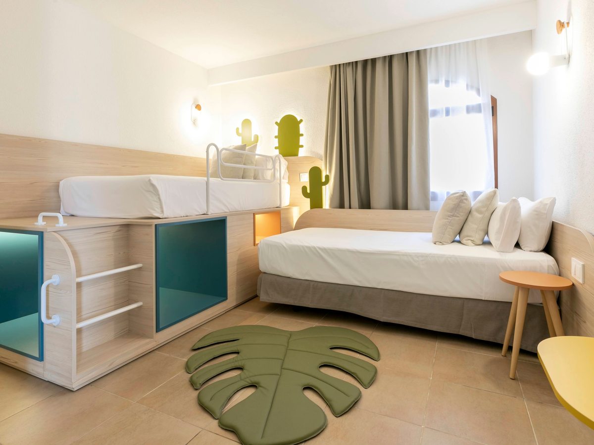 A child's bedroom with a raised single bed over a play cubby and storage, a single day bed, cactus-shaped wall lamps and a monstera-leaf rug on a tiled floor.