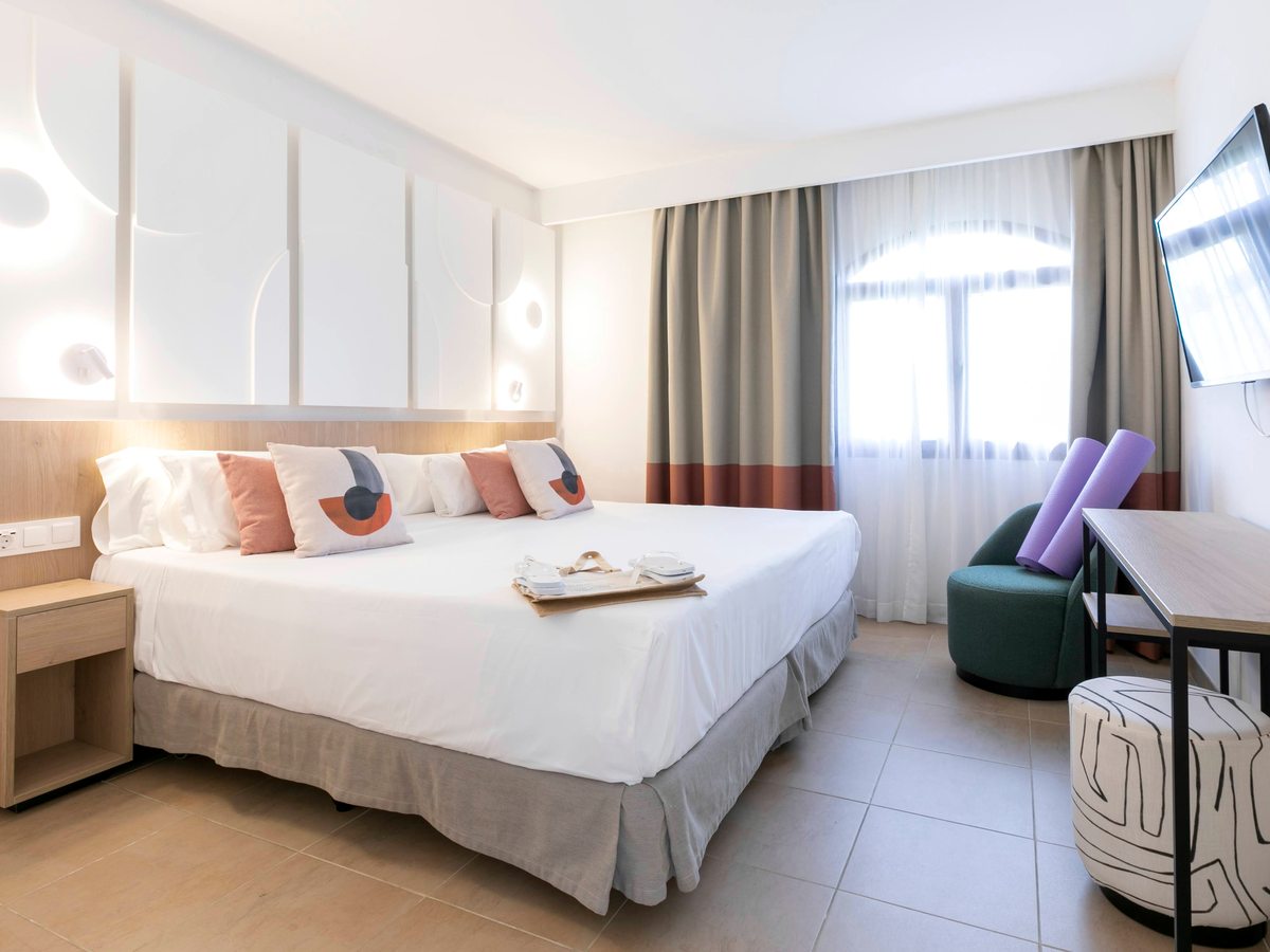 A double bedroom with a large white bed against a padded headboard, a wall-mounted TV, a desk, colourful cushions and a curtained window, in warm neutral tones.