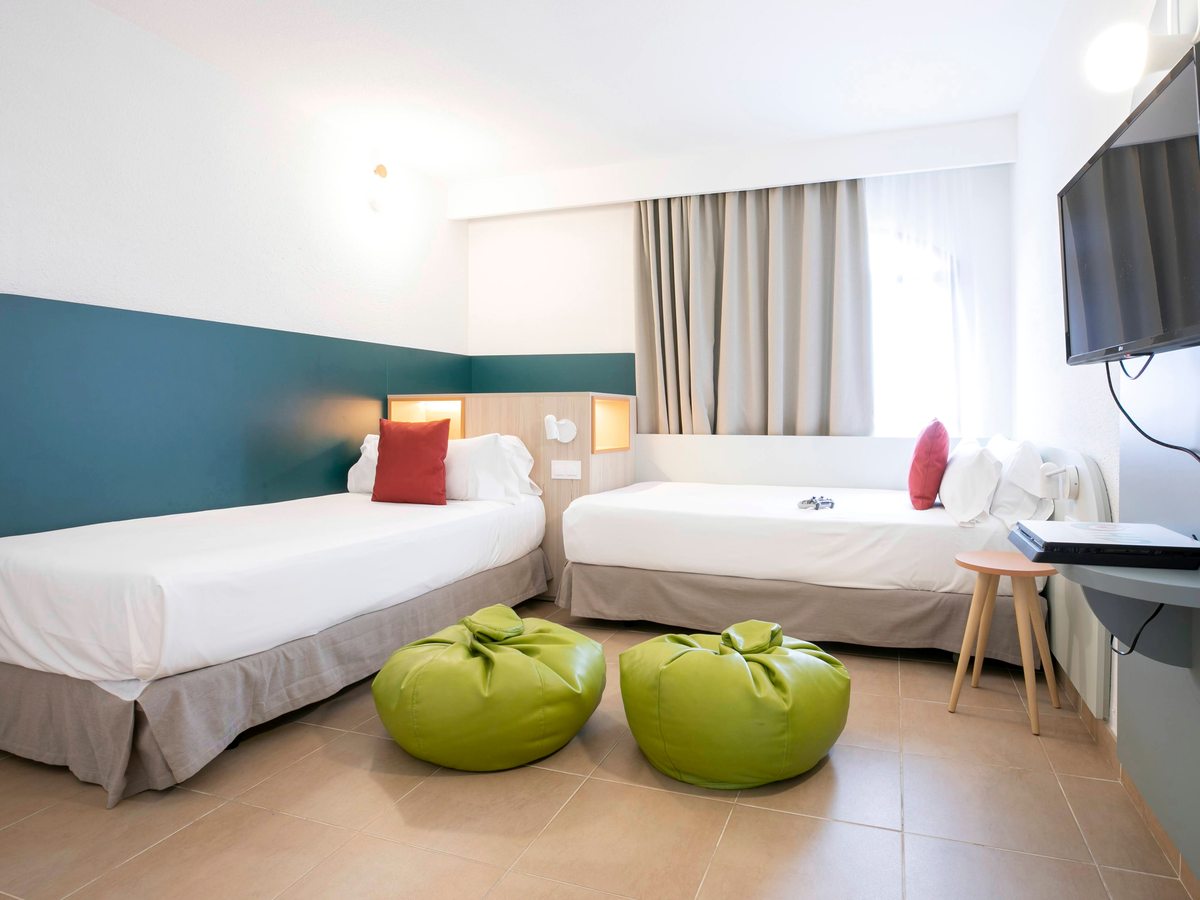 A twin bedroom with two single beds, a wall-mounted TV, lime-green beanbag seats and a teal-panelled wall on a tiled floor.