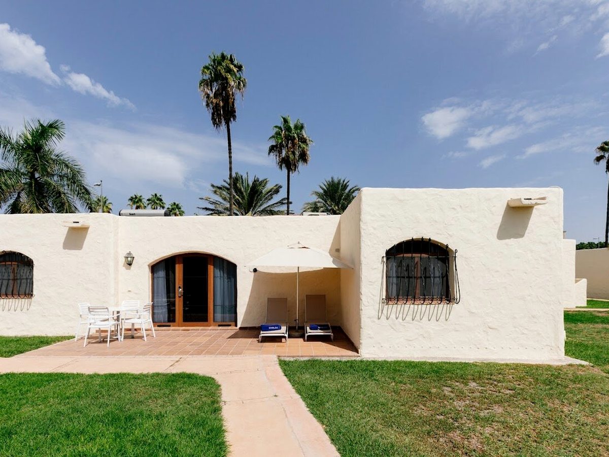 A white single-storey bungalow with an arched doorway and a small paved terrace with loungers, set on a garden lawn with palms behind under a blue sky.