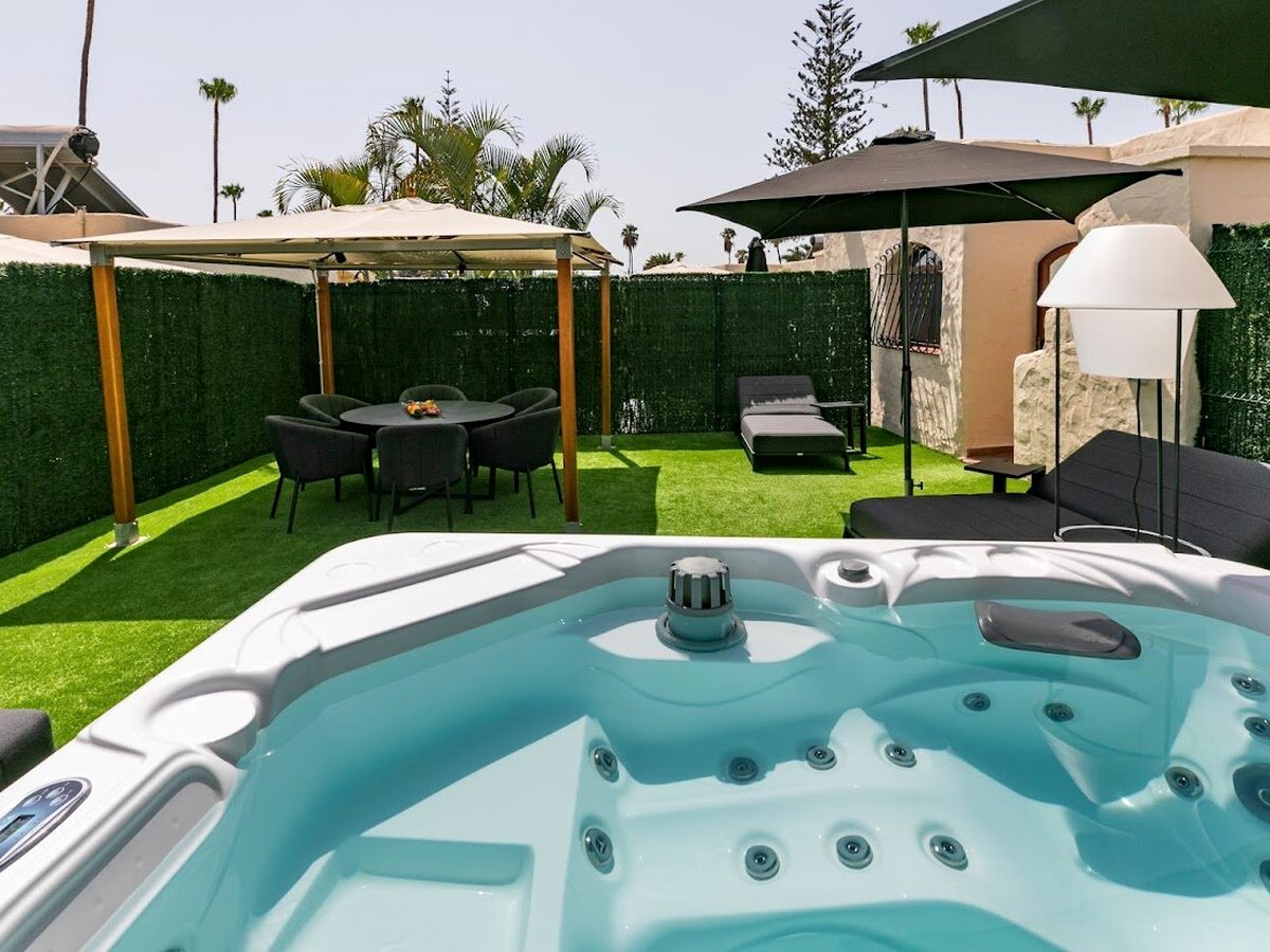 A private walled garden with a built-in jacuzzi in the foreground, a gazebo dining set, sun loungers and parasols on artificial lawn beside a bungalow, palms behind under a blue sky.