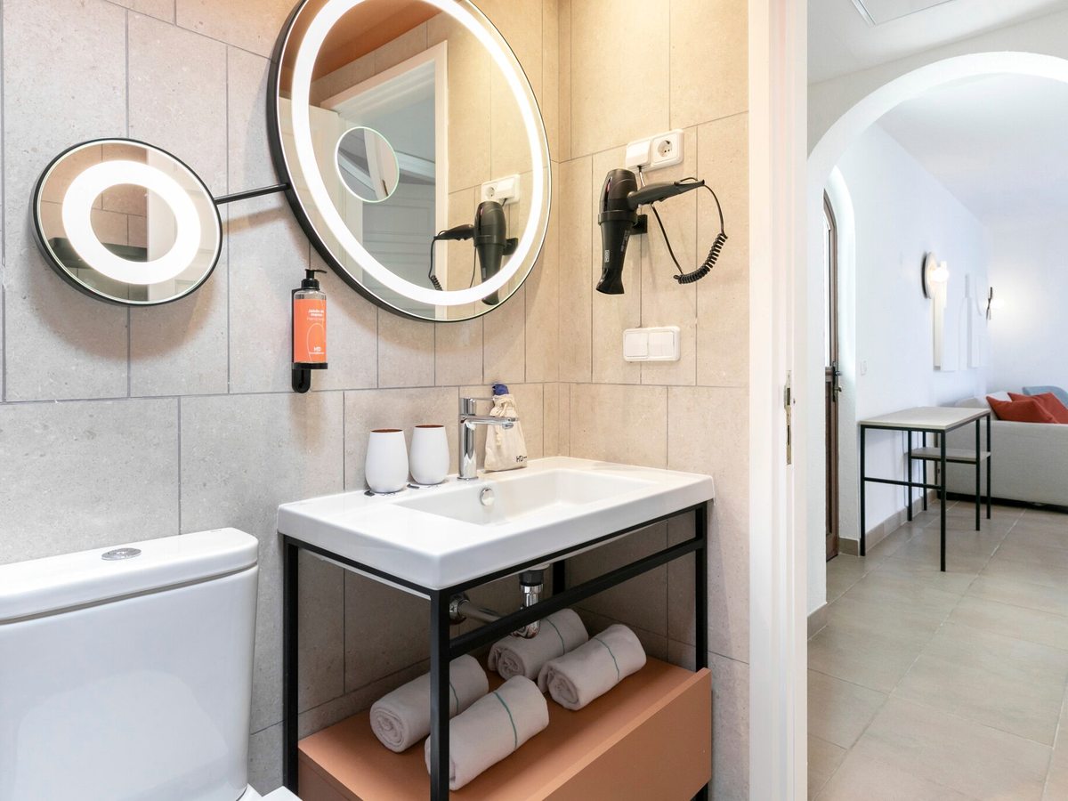 A bathroom with a round illuminated mirror over a black-framed vanity with folded towels, a toilet alongside and a doorway through to the bedroom.