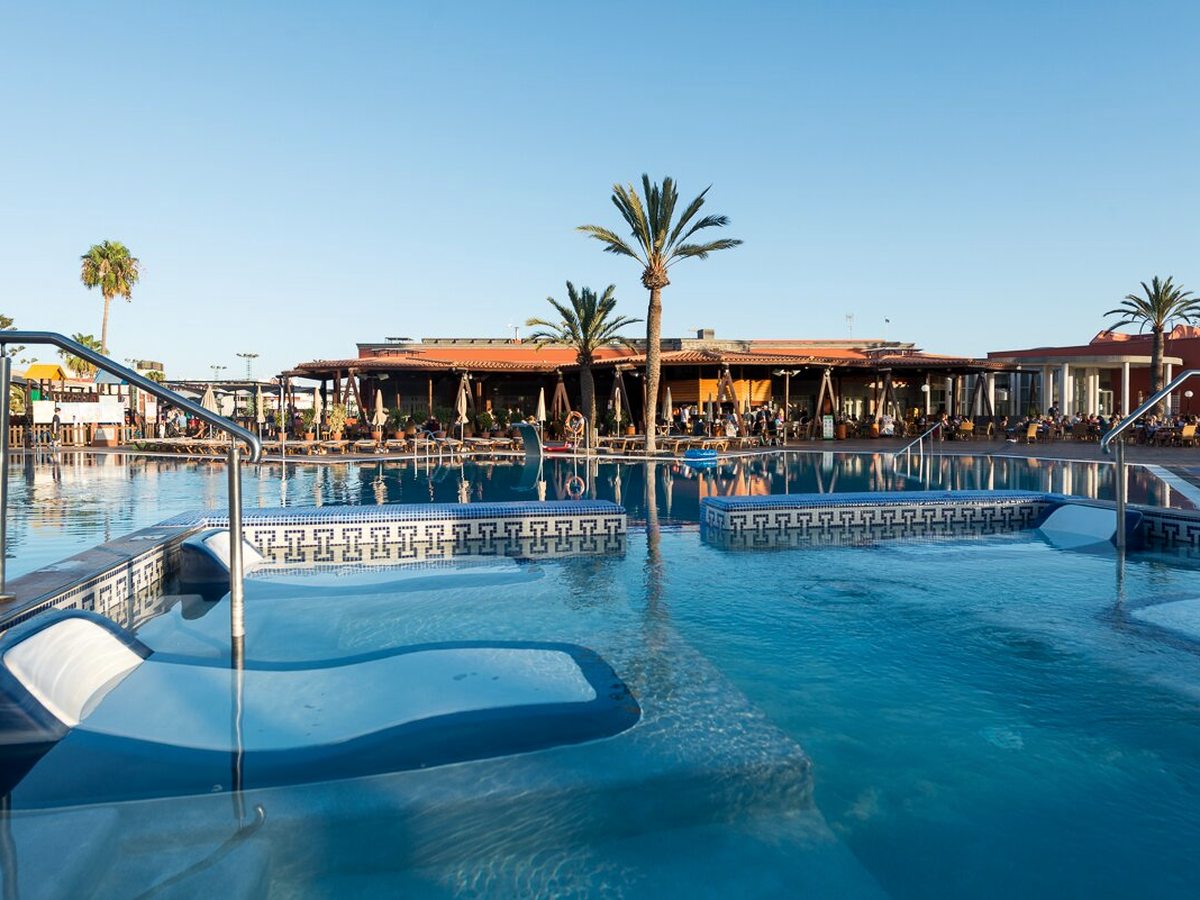 A large outdoor pool with floating loungers, sun loungers and parasols along the tiled surround and the low resort buildings and palms behind under a clear sky.