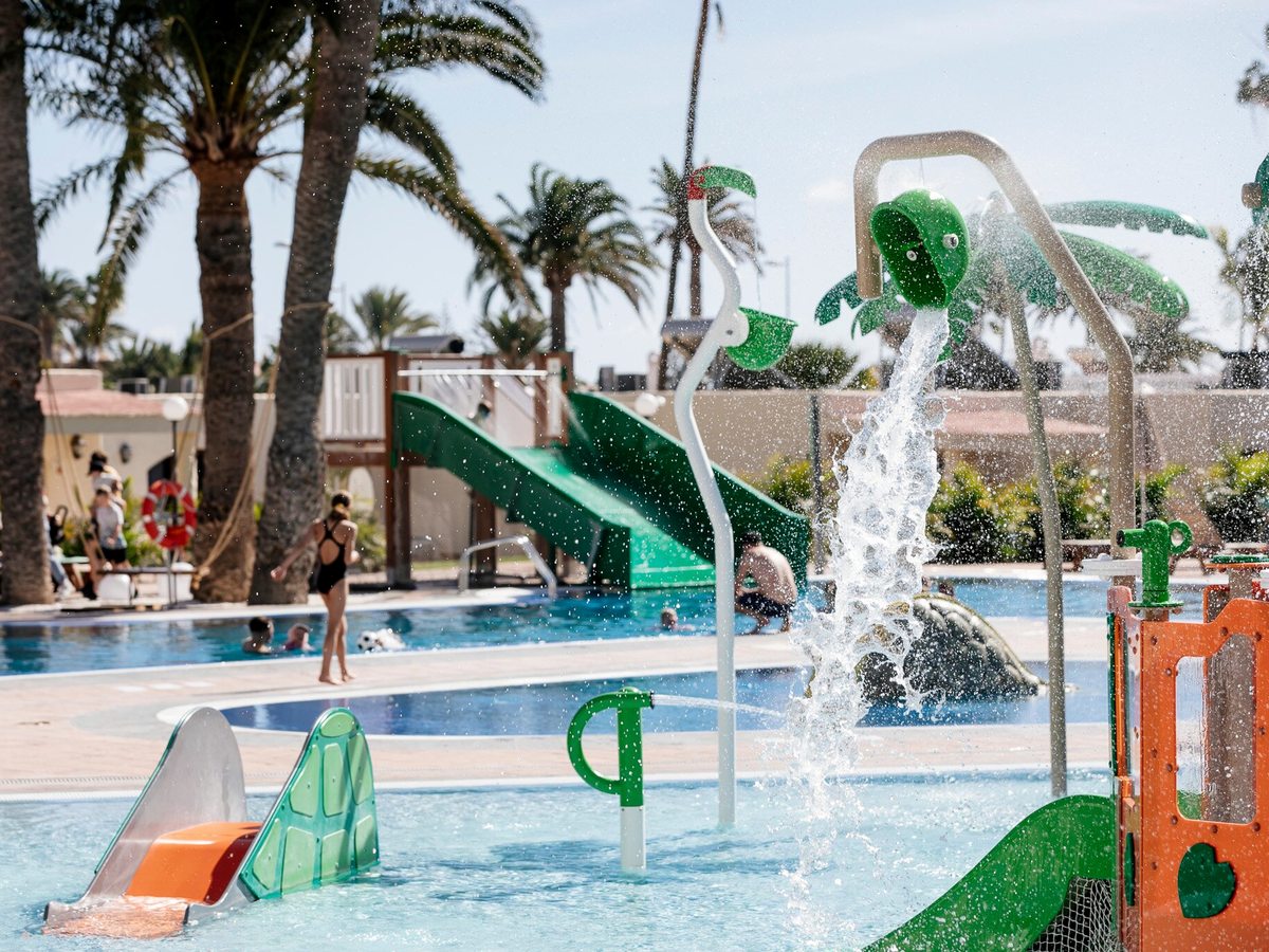 Water spouting from a raised feature and jets over a shallow splash area with green slides and palms behind under a bright sky.