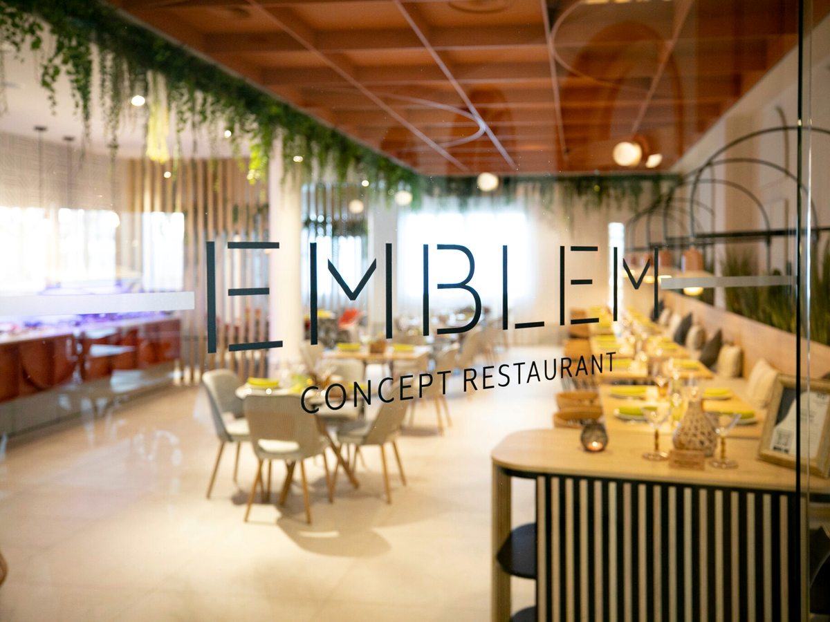 A restaurant interior with EMBLEM Concept Restaurant lettering on a glass partition, hanging greenery, a wooden ceiling and set tables beyond.