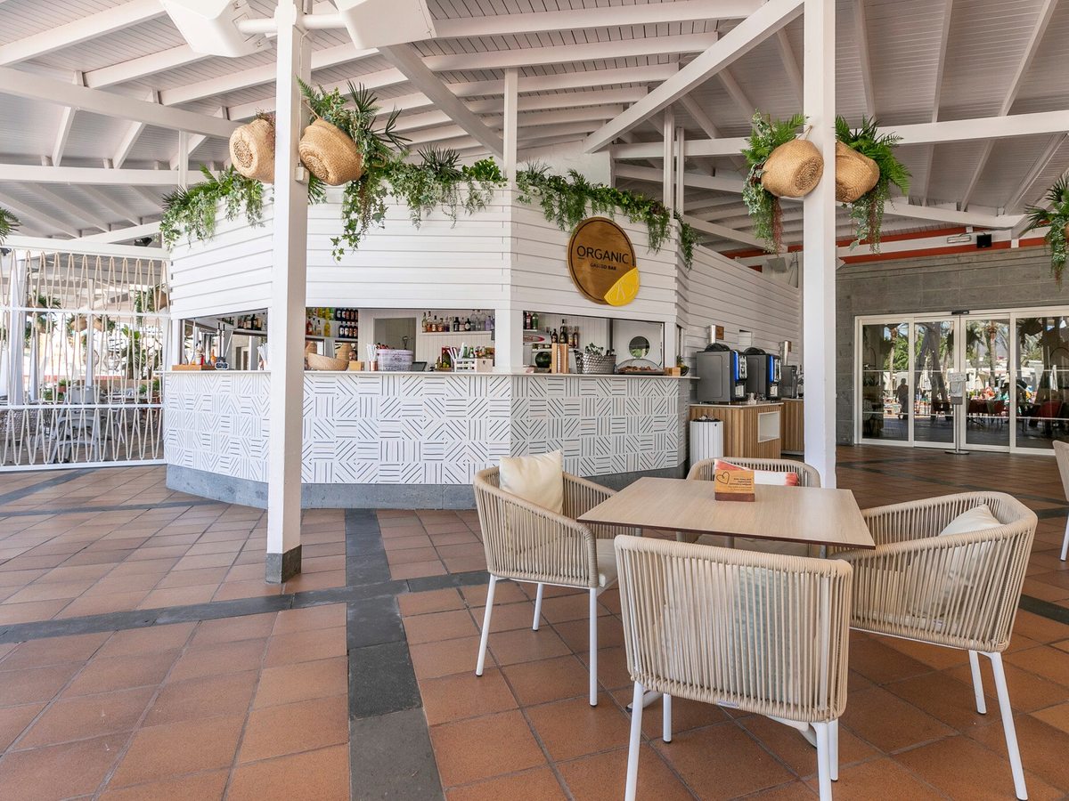 A covered open-air bar with a white pitched roof, rattan chairs and tables on a tiled terrace, greenery hanging from the beams and the bar counter to one side.