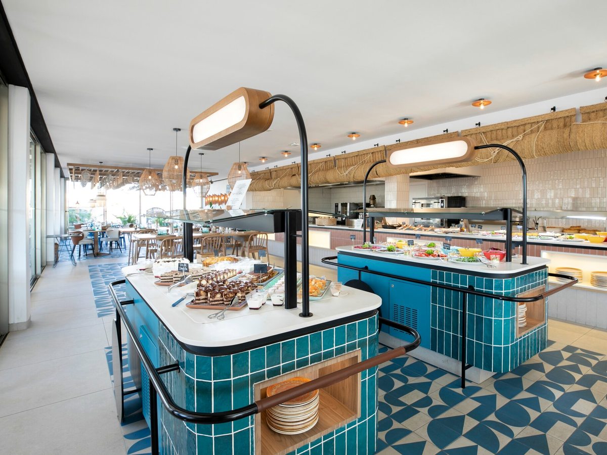 A buffet room with two teal-tiled serving islands holding desserts, salads and hot dishes under curved lamps, an open kitchen behind and dining tables to one side.