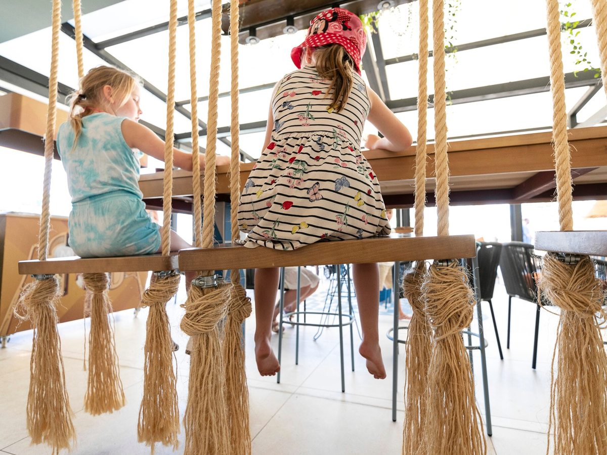 Two children in sun dresses sitting on rope swing seats at a high wooden bar counter under a pergola, with bar stools and diners behind.