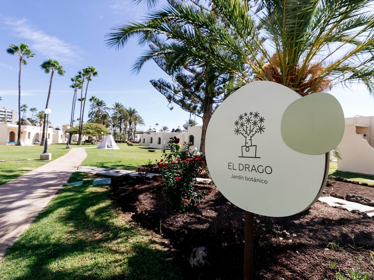 A circular sign reading El Drago beside a planted garden bed and palms, with a resort path and bungalows in the background under a bright sky.