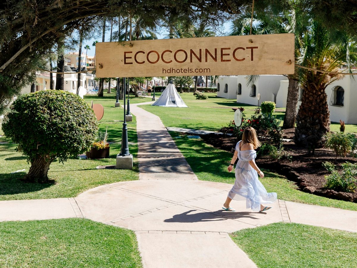 A landscaped garden walkway leading between low white bungalows, with lawns, shrubs and palms and a wooden ECOCONNECT archway over the path, a woman in a long dress walking along it.