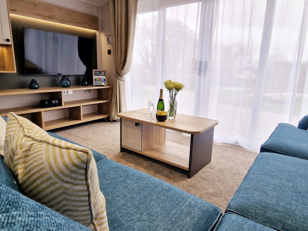 A caravan living room with a blue corner sofa, coffee table, wall-mounted TV and large glazed doors.