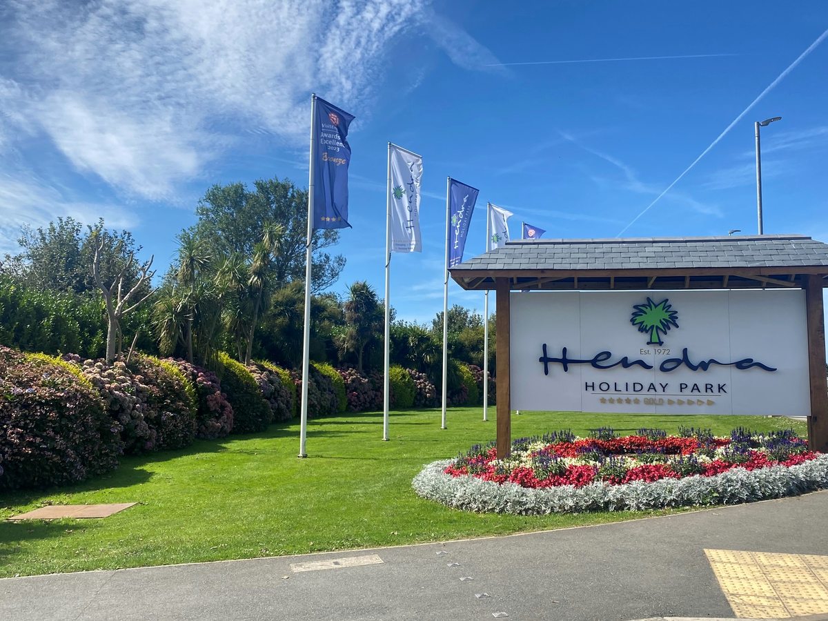 The Hendra Holiday Park entrance sign on a landscaped roadside with flagpoles and flower beds.