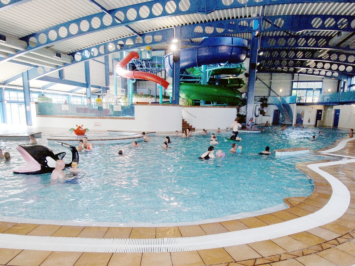 A large indoor leisure pool with a curved shallow edge, flumes behind and a high glazed roof.