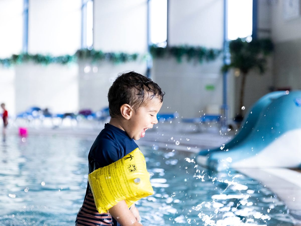 A young child in yellow armbands splashing beside a small blue slide in the shallow indoor pool.