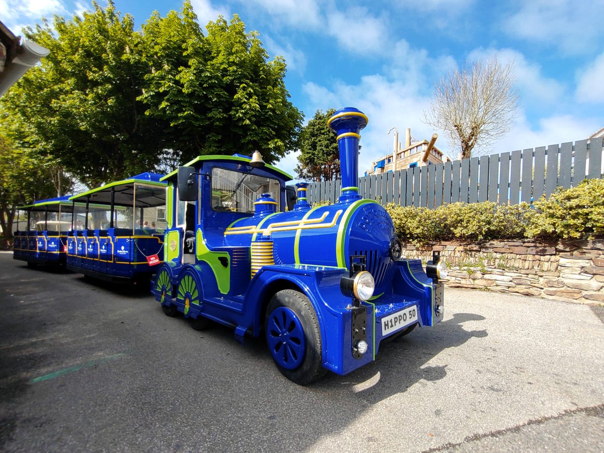The park's blue and green road train parked on a park lane beside a stone wall and hedge.