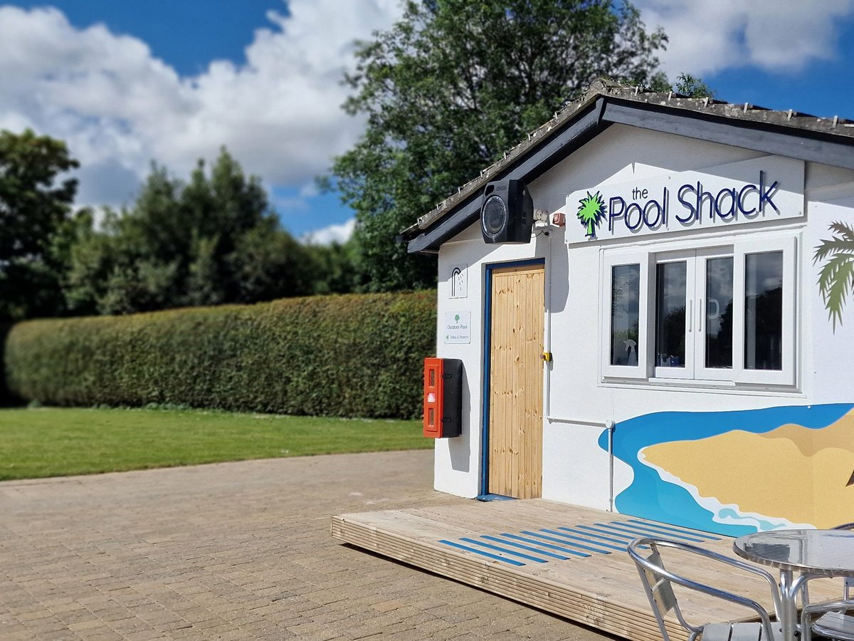 A small white poolside snack kiosk with a Pool Shack sign, tables and chairs on a paved terrace.
