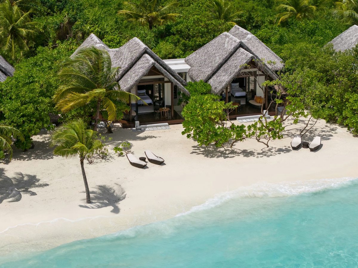 Aerial view of white sand and turquoise shallows running along the thatched beach villas and their palms.