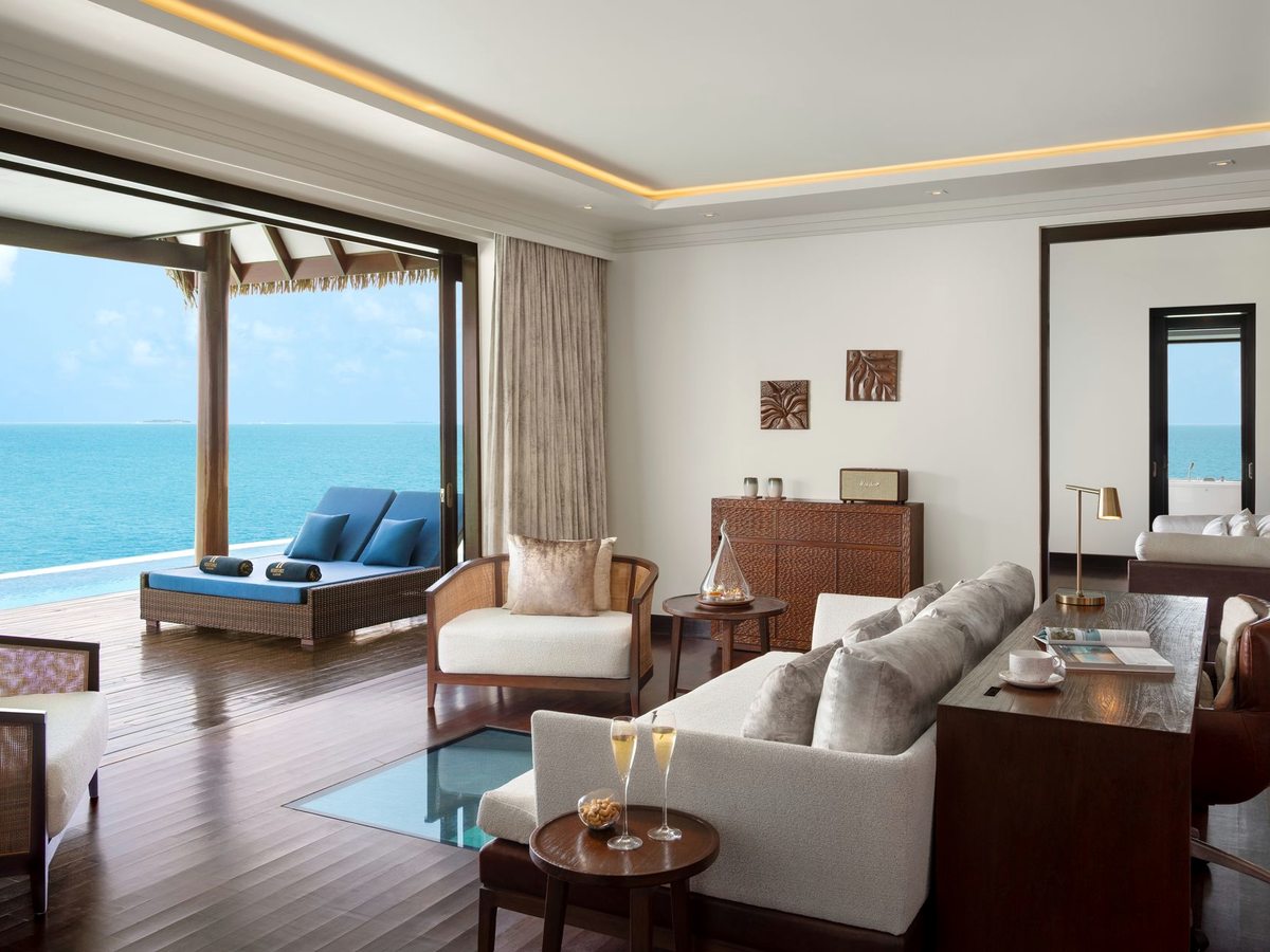 A villa living room with pale sofas, an armchair and a low table, glass doors opening to a deck and the sea.