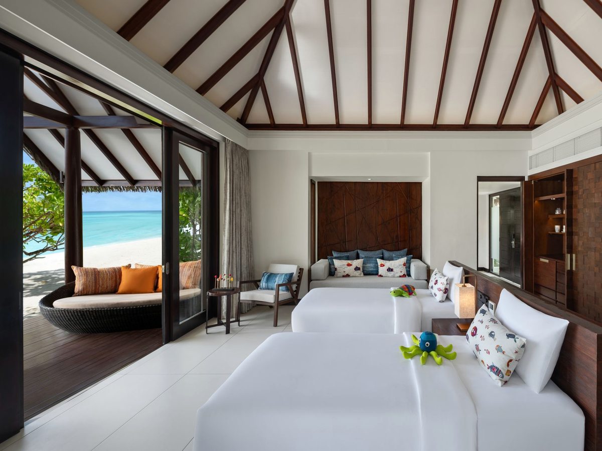A bright villa bedroom with a king bed, a curved built-in sofa and glass doors opening to the beach and sea.