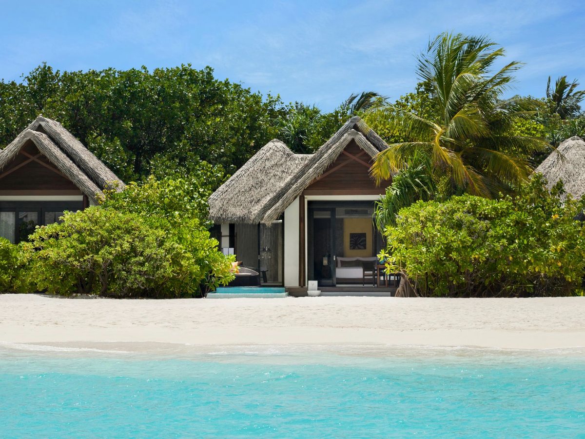 Two thatched-roof beach villas set among tropical greenery on white sand, with the turquoise lagoon in front.
