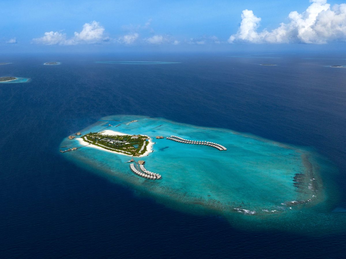 Aerial view of the whole resort island, thatched villas among dense palms, ringed by white sand and a turquoise reef in open ocean.